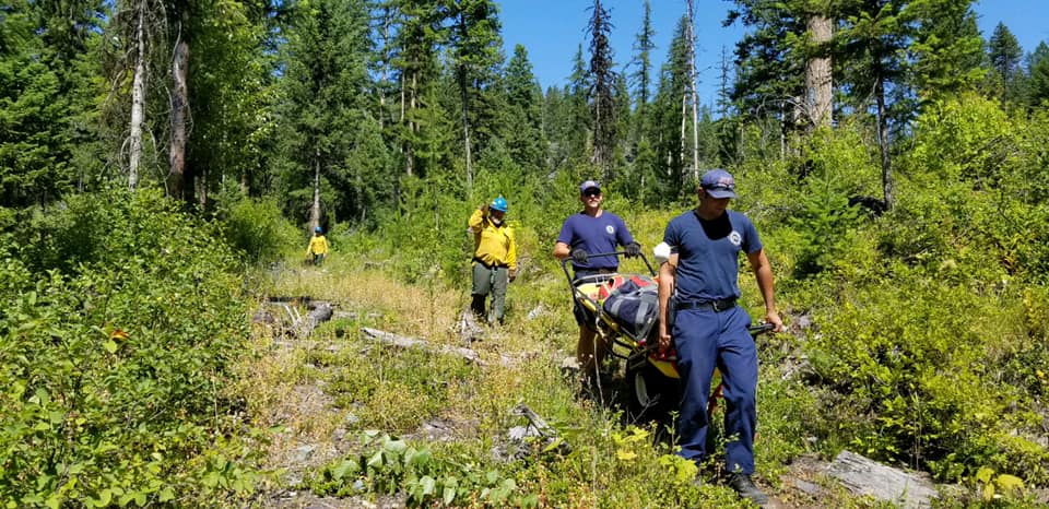 two firefighters carrying a hurt patient on the stretcher with a person walking behind them