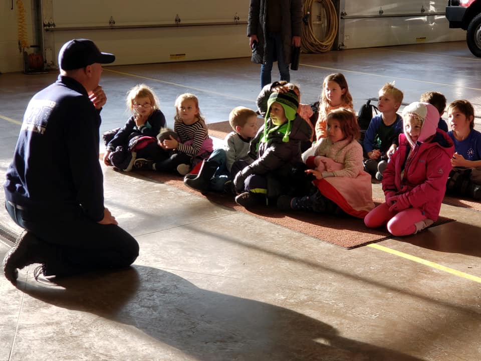 A firefighter kneeling on the ground talking to school kids sitting on the ground